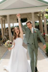 Ein fröhliches Paar feiert seine Hochzeit. Die Braut in einem weißen Kleid hält einen Blumenstrauß und streckt triumphierend den Arm in die Höhe. Der Bräutigam in einem grünen Anzug teilt den Moment mit ihr. Sie schreiten einen Gang im Freien unter einem Pavillon entlang.