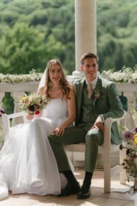 Eine Braut in einem weißen Kleid und ein Bräutigam in einem grünen Anzug sitzen lächelnd auf einer Bank im Freien. Die Braut hält einen bunten Blumenstrauß in der Hand. Sie sind von üppigem Grün und Blumenarrangements umgeben.