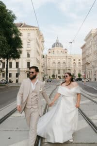 Ein Paar im Hochzeitskleid geht Hand in Hand eine Stadtstraße entlang. Der Bräutigam trägt einen beigen Anzug und eine Sonnenbrille, während die Braut ein weißes schulterfreies Kleid und eine Sonnenbrille trägt. Im Hintergrund sind historische Gebäude und Straßenbahnschienen zu sehen.
