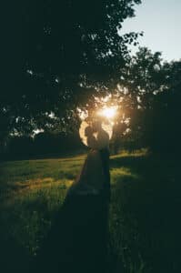 Ein Paar steht bei Sonnenuntergang auf einer Wiese, die Sonne wirft ihre Schatten auf sie. Sie umarmen sich unter einem großen Sonnenschirm, umgeben von Bäumen, durch die sanftes Licht fällt.