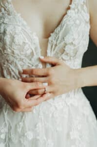 Eine Braut in einem detailreichen Spitzenhochzeitskleid hält sanft ihre Hände zusammen und betont einen Ring an ihrem Finger. Das Kleid ist mit Blumenstickereien und einem tiefen Ausschnitt versehen.