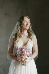 Eine Braut in einem Spitzenhochzeitskleid hält einen bunten Blumenstrauß. Ihr langes, welliges Haar ist teilweise von einem Schleier bedeckt und sie lächelt sanft und schaut zur Seite. Der Hintergrund ist eine schlichte, dunkle Wand, die der Szene eine heitere und elegante Atmosphäre verleiht.