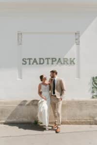 Braut und Bräutigam stehen vor einer weißen Wand mit der Aufschrift „Stadtpark“. Die Braut trägt eine Sonnenbrille und hält ihr Kleid, während der Bräutigam einen beigen Anzug trägt. Beide lächeln sich im Sonnenlicht an.