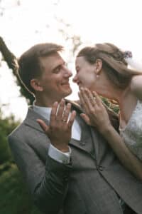 Ein glückliches Paar steht an seinem Hochzeitstag im Freien und schaut sich liebevoll an. Der Bräutigam trägt einen grauen Anzug und die Braut ein weißes Kleid. Sie zeigen lächelnd ihre Eheringe, umgeben von Grün und sanftem Sonnenlicht.