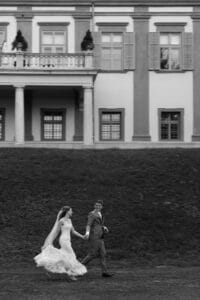 Eine Braut und ein Bräutigam laufen fröhlich Hand in Hand vor einem großen, eleganten Gebäude mit hohen Fenstern und Säulen. Die Braut trägt ein langes Kleid mit Schleier, während der Bräutigam einen Anzug trägt. Die Szene vermittelt ein Gefühl von Glück und Feierlichkeit.