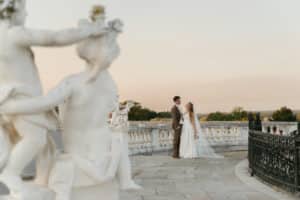 Braut und Bräutigam stehen auf einer Steinterrasse und blicken sich liebevoll an. Statuen und ein verziertes Geländer rahmen die Szene ein. Im Hintergrund sind ein klarer Himmel und Bäume in der Ferne zu sehen.