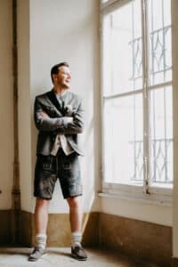 Ein Mann in traditioneller bayerischer Kleidung, darunter eine graue Jacke, ein weißes Hemd, knielange Ledershorts und Wollsocken mit Schuhen, steht mit verschränkten Armen an einem großen Fenster, lächelt und blickt in einen hellen Raum hinaus.