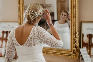 Eine Braut in einem Spitzenhochzeitskleid und einem Haarschmuck mit Blumenmuster richtet ihr Haar, während sie in einen großen, verzierten Spiegel blickt. Sie lächelt ihr Spiegelbild sanft an und schafft so eine elegante und heitere Atmosphäre.