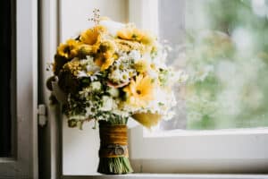 Ein Strauß gelber und weißer Blumen, darunter Gänseblümchen und Chrysanthemen, ist in gelben Stoff eingewickelt und mit einer Uhr versehen. Er steht auf einer Fensterbank und wird von sanftem, natürlichem Licht erhellt.