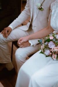 Ein sitzendes Paar, das Händchen hält. Der Mann trägt einen beigen Anzug mit einer Ansteckblume und die Frau trägt ein weißes Kleid und hält einen Strauß rosa und weißer Blumen. Sie sitzen auf Holzstühlen, neben ihnen ist eine weitere Person teilweise sichtbar.