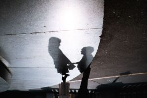 Der Schatten zweier Menschen, möglicherweise Kinder, die sich an den Händen halten, wird auf einen strukturierten Bürgersteig projiziert. Die Lichtquelle wirft ihre Silhouetten so, dass die Figuren langgestreckt erscheinen.