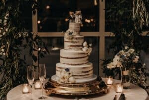 Eine dreistöckige, nackte Hochzeitstorte, geschmückt mit weißen und blauen Blumen und mit Bärenfiguren obendrauf, steht auf einem goldenen Tablett. Um sie herum sind Kerzen, zwei Champagnergläser und ein grünes Blattwerkdekor angebracht, was eine romantische Atmosphäre schafft.
