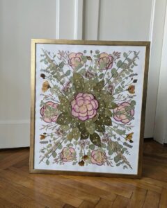 Ein gerahmtes Blumenkunstwerk mit einer symmetrischen Anordnung von rosa und grünen Blumen mit Blättern vor einem weißen Hintergrund. Der Rahmen hat eine goldene Oberfläche und steht auf einem Holzboden, an eine Wand gelehnt.
