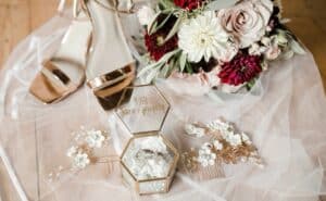 Brautaccessoires auf Tüllstoff. Enthält metallische High Heels, Blumenhaarnadeln und einen Strauß rosa und roter Blumen mit Grün. Eine sechseckige Glasbox enthält einen Ring mit der Aufschrift „Love Always“.