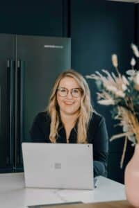 Eine Frau mit langen Haaren und Brille sitzt an einem Tisch und lächelt auf einen Laptop. Sie befindet sich in einer modernen Küche mit dunklen Schränken und einer Vase mit getrockneten Blumen in der Nähe.