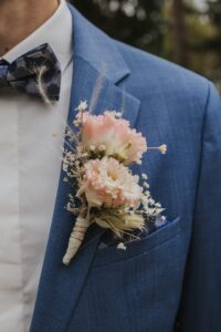 Eine Nahaufnahme einer Person, die einen blauen Anzug und eine Fliege mit Blumenmuster trägt. An der Anzugjacke ist eine Ansteckblume mit rosa Blumen und kleinen weißen Blüten befestigt.