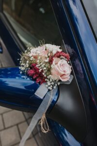 Ein Strauß rosa Rosen, weißem Schleierkraut und roten Blumen ist mit einem Band am Seitenspiegel eines blauen Autos befestigt. Das Auto steht auf einer gepflasterten Fläche und spiegelt den klaren Himmel wider.