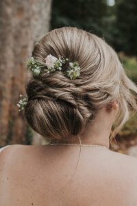 Das Bild zeigt das Haar einer Frau, das zu einer eleganten Hochsteckfrisur mit weichen Locken gestylt ist. Es ist mit kleinen weißen Blumen und Grünpflanzen geschmückt. Im Hintergrund sind ein Baumstamm und verschwommenes Grün zu sehen, was eine Umgebung im Freien suggeriert.