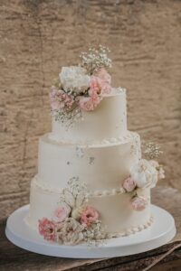 Eine dreistöckige weiße Hochzeitstorte, geschmückt mit rosa und weißen Blumen, darunter Rosen und Schleierkraut, vor einem rustikalen Steinhintergrund.