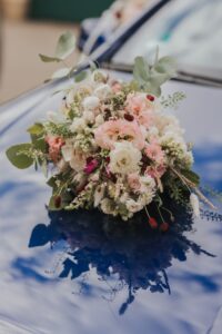 Ein Strauß rosa, weißer und grüner Blumen liegt auf der Motorhaube eines blauen Autos. Auf der Oberfläche des Autos spiegeln sich Himmel und Wolken.