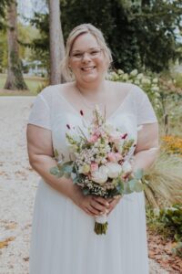Eine lächelnde Braut in einem weißen Brautkleid und mit Brille steht draußen und hält einen Strauß rosa und weißer Blumen in der Hand. Im Hintergrund sind Bäume und Grün zu sehen.