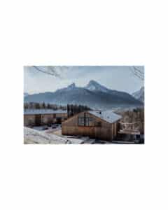 Schneebedeckte Holzgebäude zwischen Bäumen, mit einer majestätischen Bergkette im Hintergrund. Der Himmel ist klar und die Szene ist ruhig und heiter und stellt eine Winterlandschaft dar.