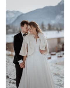 Ein Paar in Hochzeitskleidung steht dicht beieinander und lächelt sanft, umgeben von einer verschneiten Landschaft. Die Braut trägt ein weißes Kleid mit einem Pelzschal und der Bräutigam einen dunklen Anzug. Im Hintergrund sind Berge und Bäume zu sehen.