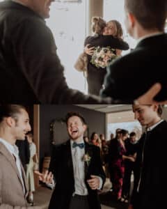 Bei einem Treffen umarmt eine Frau einen Freund, während sie einen Blumenstrauß hält. Ihr folgt ein fröhlicher Mann im Anzug, der mit zwei anderen lacht, einer trägt eine braune Jacke und der andere einen dunklen Blazer. Andere sind im Hintergrund zu sehen.