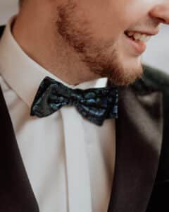 Eine Person im Smoking mit einer glänzenden, dunklen Fliege mit Blumenmuster und einem weißen Hemd lächelt. Der Fokus liegt auf der Fliege und dem unteren Teil des Gesichts, wodurch ein freudiger Ausdruck eingefangen wird.