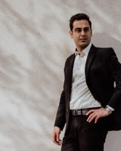 Ein Mann in schwarzem Anzug und weißem Hemd steht selbstbewusst mit einer Hand auf der Hüfte vor einer strukturierten, hellen Wand. Er hat dunkles Haar, eine Brille und einen entspannten Gesichtsausdruck. Weiche Schatten werden auf die Wand geworfen.