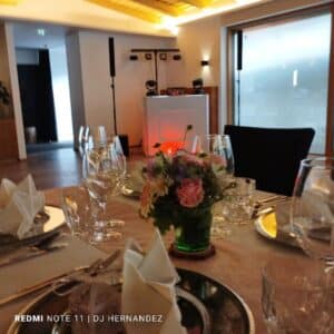 Ein gedeckter Tisch bei einem Bankett mit gefalteten Servietten, Glaswaren und einem Blumenschmuck. Im Hintergrund steht ein DJ-Pult mit Lautsprechern und Beleuchtungsanlage. Der Raum hat ein modernes Ambiente mit gedämpfter Beleuchtung.