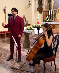Ein Mann im roten Anzug singt, während er neben einer sitzenden Frau steht, die in einer Kirche Cello spielt. Die Kulisse umfasst religiöse Statuen, Blumenarrangements und Kerzen.