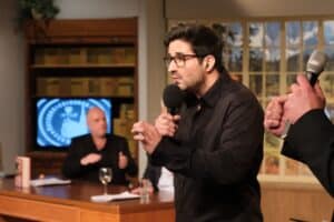 Ein Mann mit Brille und schwarzem Hemd spricht in ein Mikrofon. Eine andere Person hält ein Mikrofon. Im Hintergrund sitzen zwei Personen an einem Schreibtisch. Ein Buch, ein Glas und ein Fernsehbildschirm sind zu sehen.