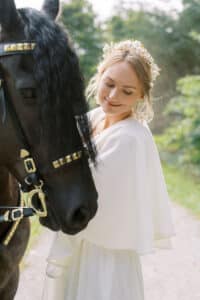 Eine Frau in einem weißen Kleid und einem Blumenkopfschmuck lächelt sanft neben einem schwarzen Pferd mit verzierten Zügeln. Sie stehen auf einem von Grün umgebenen Weg.