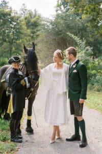 Eine Braut in einem weißen Kleid und ein Bräutigam in traditioneller Kleidung stehen neben einem schwarzen Pferd auf einem Weg. Eine Person in einem historischen Kostüm hält die Zügel des Pferdes. Im Hintergrund sind sie von Bäumen und Grün umgeben.