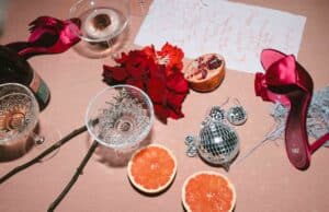 Eine festliche Tischszene mit zwei roten Stöckelschuhen, verstreuten roten Blumen, einem halben Granatapfel, Glaswaren, rosa Grapefruithälften, Discokugel-Ornamenten, einem dekorativen Zweig und einer handgeschriebenen Notiz auf einer hellen Tischdecke.