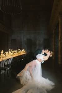 Ein Paar tanzt in einer eleganten, schwach beleuchteten Umgebung, wobei der Mann die Frau, die ein fließendes weißes Kleid trägt, in den Tanz taucht. Im Hintergrund steht ein langer Tisch, der mit Blumen und Kerzen dekoriert ist und zur romantischen Atmosphäre beiträgt.