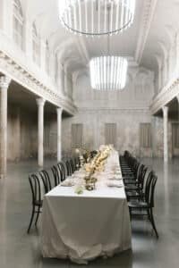 Eleganter Bankettsaal mit langem Esstisch, der für eine formelle Veranstaltung gedeckt ist. Der Tisch ist mit weißer Tischwäsche, Blumendekorationen und Geschirr geschmückt. Schwarze Stühle stehen auf beiden Seiten. Große Kronleuchter hängen von der hohen, verzierten Decke und erhellen den Raum.