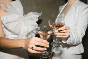 Zwei Personen in eleganter weißer Kleidung stoßen mit Champagnergläsern an, die mit einem hellrosa Getränk gefüllt sind. Eine Person trägt ein Kleid mit einer großen Stoffschleife. Die andere trägt einen Anzug. Der Hintergrund ist eine neutrale, strukturierte Wand.