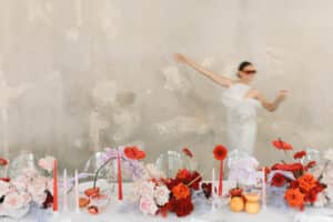Eine Frau in einem weißen Kleid und roter Sonnenbrille hält ein Getränk in der Hand, das Bild ist verschwommen in der Bewegung. Der Tisch davor ist mit roten, rosa und weißen Blumen, Kerzen und Zitrusfrüchten geschmückt. Der Hintergrund ist eine strukturierte graue Wand.