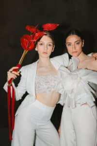 Zwei Frauen in eleganten weißen Outfits posieren selbstbewusst. Eine hält rote Anthurienblüten, die andere ein großes weißes Blumenaccessoire. Der dunkle Hintergrund kontrastiert mit ihrer hellen Kleidung und hebt die Blumen und Details hervor.