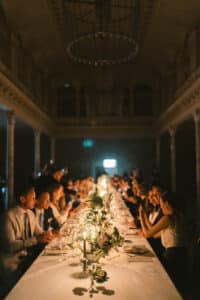 Elegante Dinnerszene in einem schwach beleuchteten, großen Saal mit einem langen, dekorierten Tisch. Die Gäste in formeller Kleidung unterhalten sich. Blumenschmuck und stimmungsvolles Licht schaffen eine intime Atmosphäre.