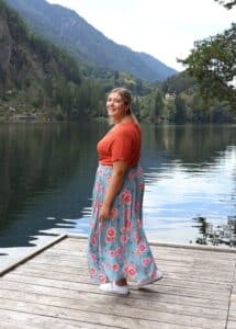 Eine Person in einem orangefarbenen Oberteil und einem geblümten Rock steht auf einem Holzsteg an einem ruhigen See, umgeben von bewaldeten Hügeln und Bergen. Der Himmel ist klar und das ruhige Wasser spiegelt die Landschaft wider.