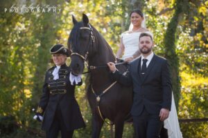Eine Frau in einem weißen Kleid sitzt lächelnd auf einem schwarzen Pferd. Ein Mann in Abendgarderobe hält die Zügel des Pferdes. Eine weitere Person in einer historischen Uniform steht neben ihnen. Sie sind im Freien, umgeben von Bäumen.