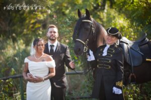 Eine Braut in einem weißen schulterfreien Kleid und ein Bräutigam in einem schwarzen Anzug stehen neben einem schwarzen Pferd. Eine Person in einer historischen Uniform mit einem Dreispitz hält die Zügel des Pferdes. Im Hintergrund sind Bäume mit grünem Laub zu sehen.