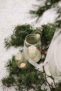 Großes Glasgefäß mit brennender Kerze auf verschneitem Boden, umgeben von Tannenzweigen. Drei kleinere Glasgefäße mit Kerzen daneben. Weiche Stoffvorhänge sind teilweise rechts sichtbar und sorgen für eine gemütliche, winterliche Atmosphäre.