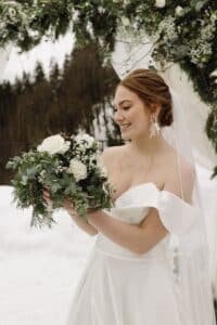 Eine Braut lächelt und hält einen Strauß weißer Rosen und Grünpflanzen in den Händen. Sie trägt ein schulterfreies weißes Kleid und einen Schleier. Im Hintergrund sind schneebedeckte Bäume zu sehen, die der Szene eine winterliche Kulisse verleihen.