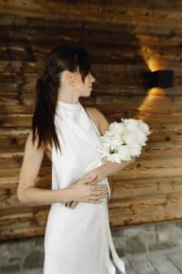 Eine Frau in einem weißen ärmellosen Kleid hält einen Strauß weißer Rosen. Sie hat langes braunes Haar, das zu einem Pferdeschwanz zurückgebunden ist. Im Hintergrund ist eine Holzwand mit dezenter Beleuchtung zu sehen.