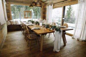 Ein rustikales Esszimmer mit einem langen Holztisch, der für eine Mahlzeit gedeckt ist und mit Grünpflanzen, Kerzen und weißer Tischwäsche geschmückt ist. Große Fenster bieten einen Blick auf den verschneiten Wald. Holzstühle und eine Bank mit Kissen bieten Sitzgelegenheiten und über dem Tisch hängt eine Hängelampe.