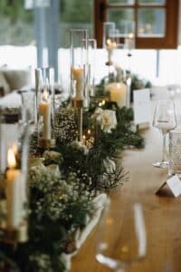 Ein für eine formelle Veranstaltung gedeckter Holztisch verfügt über große Kerzen in Glashaltern und Blumenarrangements mit weißen Blumen und Grünpflanzen. Elegantes Glasgeschirr und Namenskarten werden neben den Tafelaufsätzen platziert und sorgen für ein gehobenes Ambiente.
